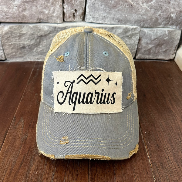 Gray trucker cap with 'Aquarius' patch on a wooden surface and stone wall background