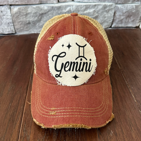 Red distressed cap with 'Gemini' design on a wooden surface and stone wall background