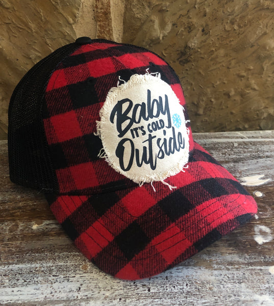Left side view of the Baby It's Cold Outside hat by BohoGroove featuring black Baby It's Cold Outside lettering and a blue snowflake graphic on a distressed off-white patch on a red and black buffalo plaid snapback trucker hat with black mesh back.