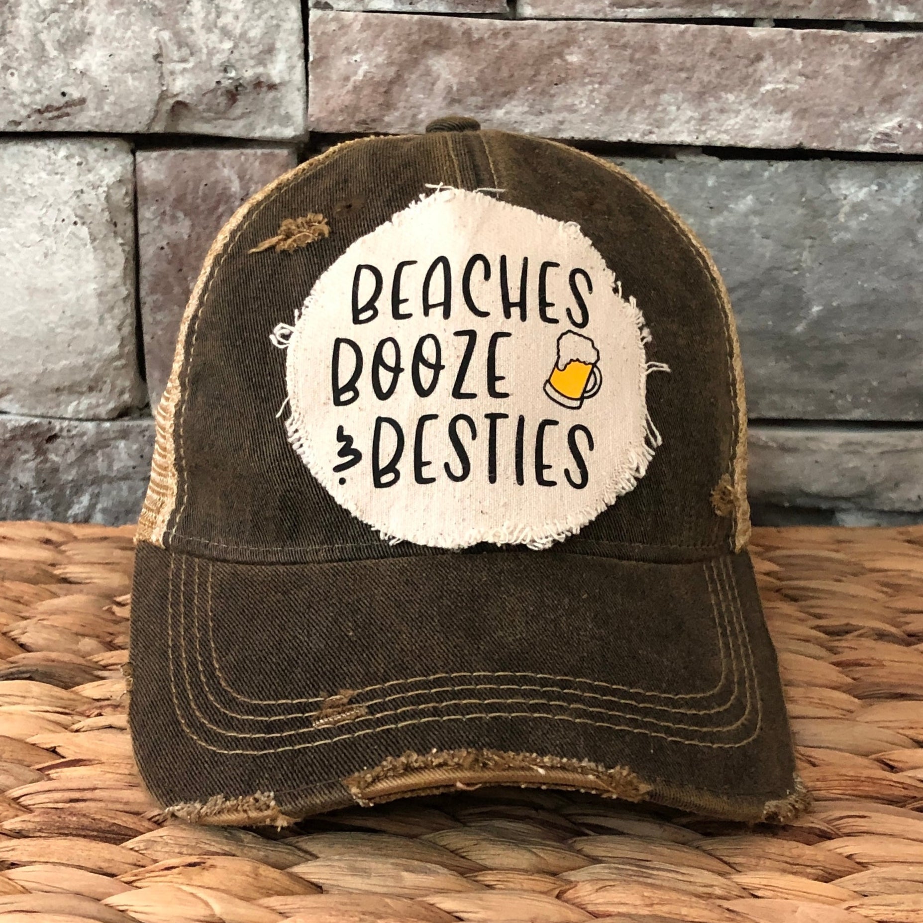 Black distressed cap with 'Beaches Booze & Besties' text on a woven surface and stone wall background