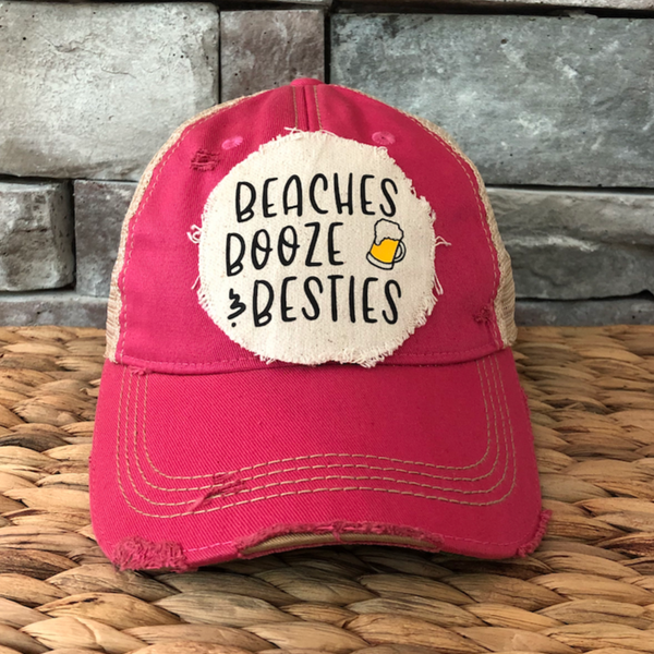 Pink cap with 'Beaches Booze Besties' text on a woven surface and stone wall background