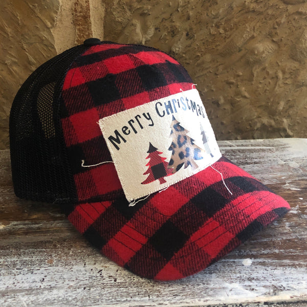 Left side view of the Buffalo Plaid Christmas Tree hat by BohoGroove featuring black Merry Christmas lettering and red, leopard print, and white Christmas tree graphics on a distressed off-white patch on a red and black buffalo plaid snapback trucker hat with black mesh back.