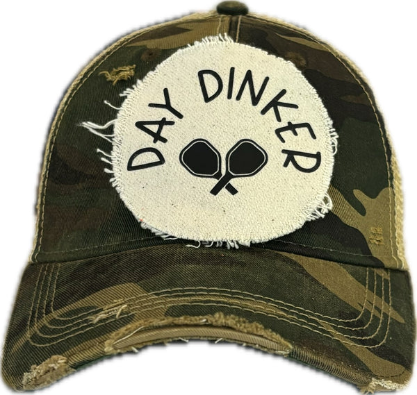 Day Dinker hat by BohoGroove featuring black Day Dinker lettering and crossed pickleball paddles graphic on a distressed off-white patch on a camo vintage distressed snapback trucker hat.