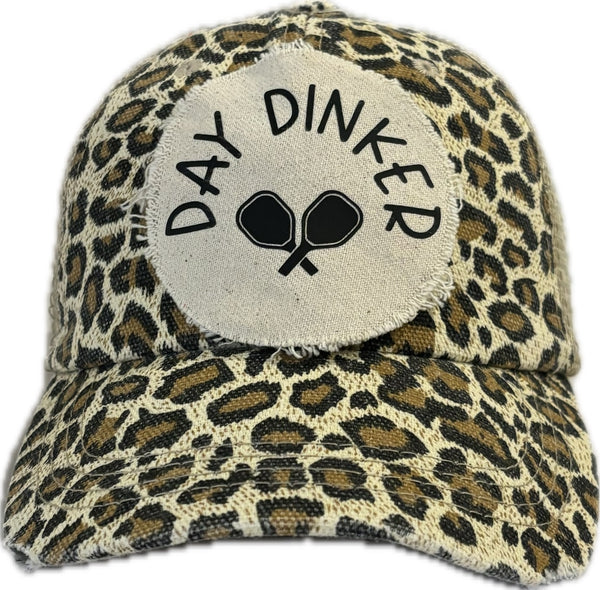 Day Dinker hat by BohoGroove featuring black Day Dinker lettering and crossed pickleball paddles graphic on a distressed off-white patch on a leopard vintage distressed snapback trucker hat.
