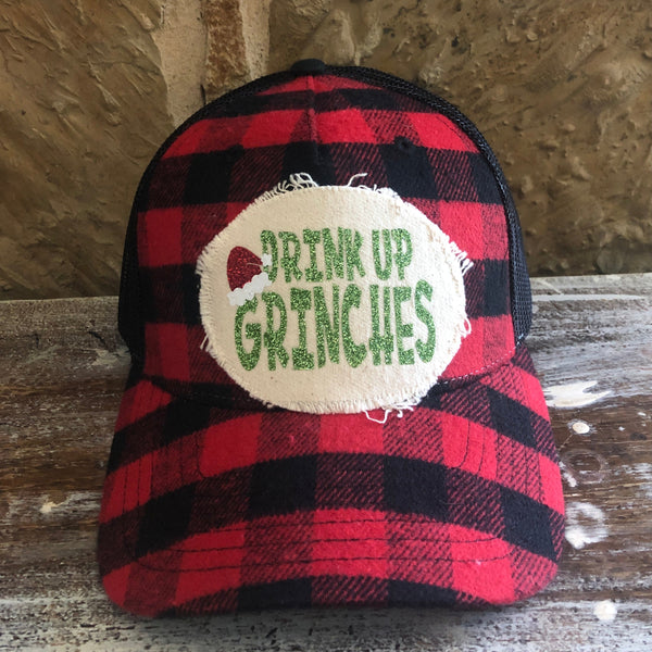 Drink Up Grinches hat by BohoGroove with green glitter Drink Up Grinches lettering and a red Santa hat graphic on a distressed off-white patch on a red and black buffalo plaid snapback trucker hat.