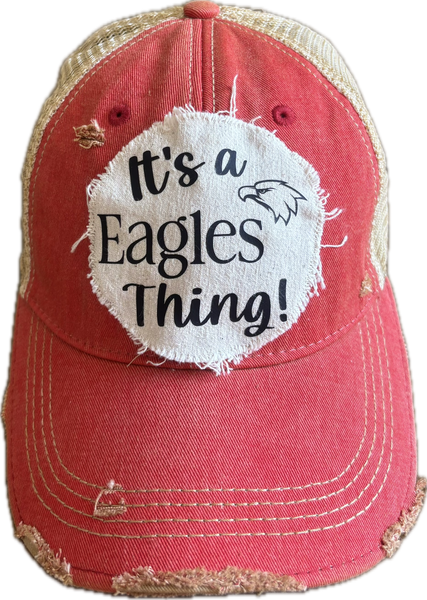 It's an Eagles Thing hat by BohoGroove featuring black lettering and an eagle graphic on a round cream frayed patch on a red vintage distressed snapback trucker hat.