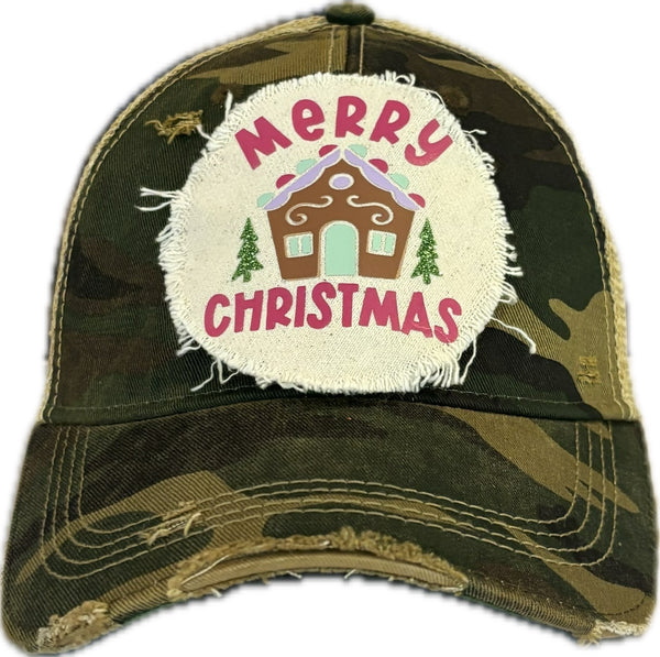 Gingerbread House hat by BohoGroove with pink Merry Christmas lettering, a gingerbread house graphic, and green trees on a distressed off-white patch on a camo vintage distressed snapback.
