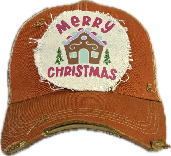 Gingerbread House hat by BohoGroove with pink Merry Christmas lettering, a gingerbread house graphic, and green trees on a distressed off-white patch on an orange vintage distressed snapback.