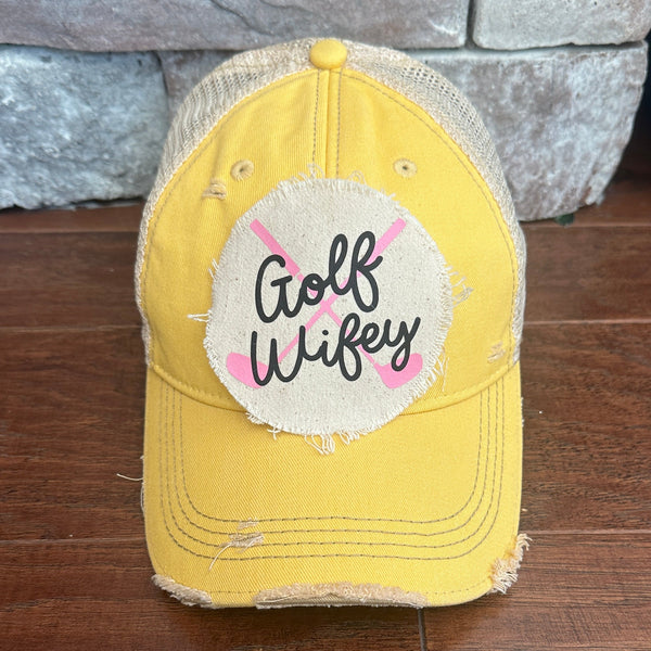 Golf Wifey hat by BohoGroove featuring black Golf Wifey lettering and pink crossed golf clubs on a yellow vintage distressed snapback hat