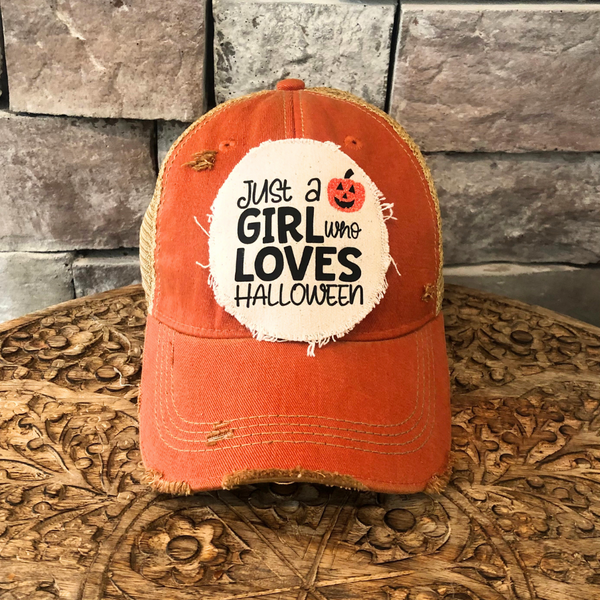 Just a Girl Who Loves Halloween hat by BohoGroove with black wording and a jack-o'-lantern graphic on a distressed off-white patch on an orange vintage distressed snapback trucker hat.