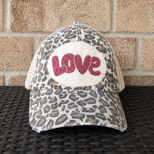 Love hat by BohoGroove with pink glitter LOVE lettering on a distressed off-white patch on a leopard vintage distressed snapback trucker hat.