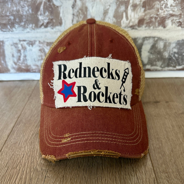 Rednecks And Rockets hat by BohoGroove with black lettering, red and blue star graphic, and firework rocket graphic on distressed off-white patch on brick red vintage distressed snapback.