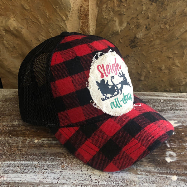 Left side view of the Sleigh All Day hat by BohoGroove featuring red Sleigh lettering, green all-day lettering, and black Santa sleigh and reindeer graphics on a distressed off-white patch on a red and black buffalo plaid snapback trucker hat with black mesh back.