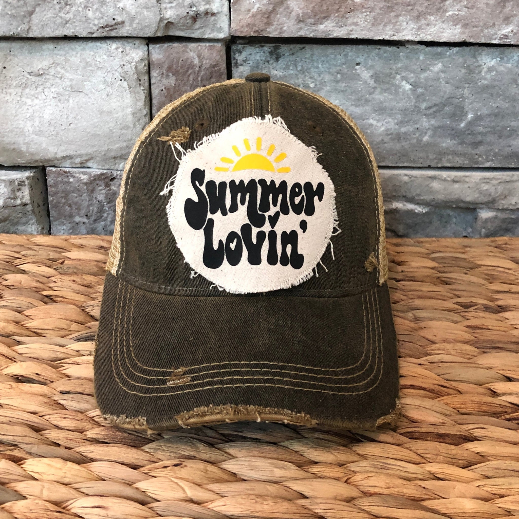Black distressed cap with 'Summer Lovin'' text on a woven surface and stone wall background