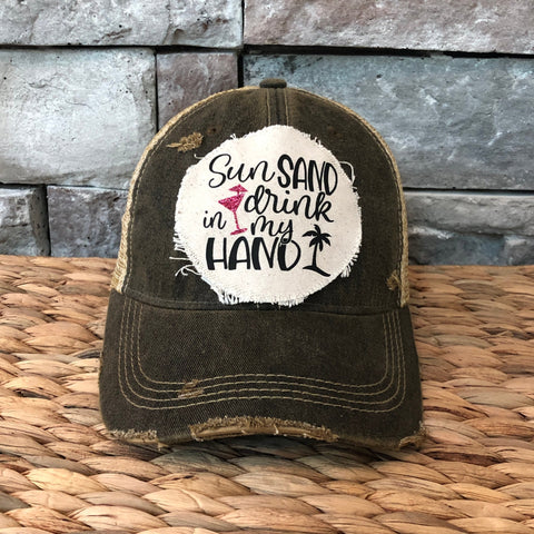 Distressed black cap with 'Sun, Sand, Drink in my Hand' text on a woven surface with stone wall background