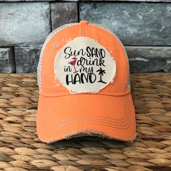 Orange cap with a patch that reads 'Sun, Sand, Drink in My Hand' on a woven surface with stone wall background.