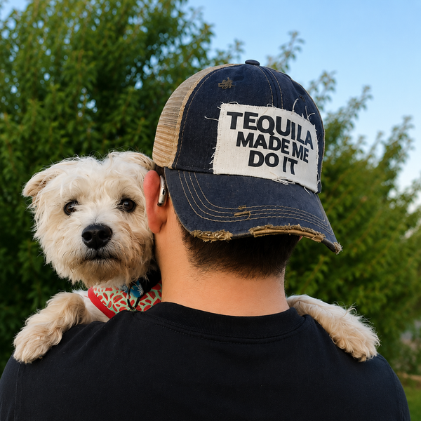 Back view of a man holding a white dog outdoors while wearing a navy vintage distressed snapback trucker hat by BohoGroove featuring a distressed off-white patch with black Tequila Made Me Do It lettering.