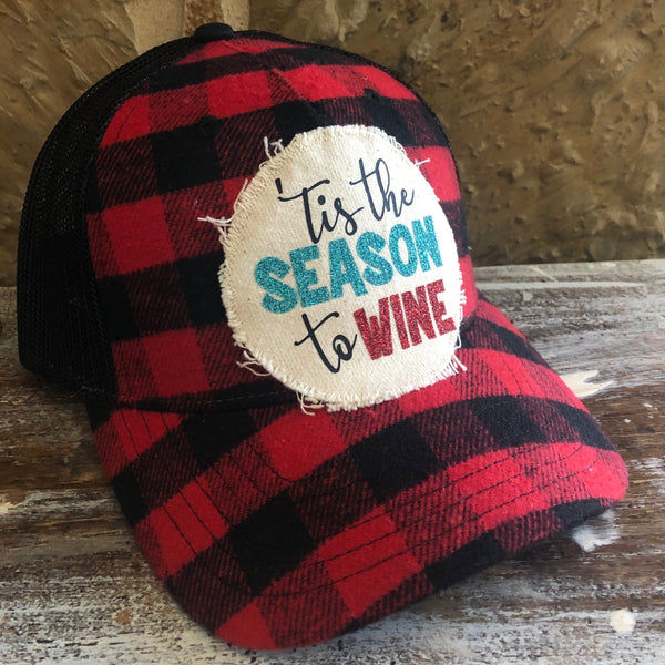 Left side view of the Tis the Season to Wine hat by BohoGroove featuring black Tis the wording, teal glitter Season lettering, and red glitter Wine lettering on a distressed off-white patch on a red and black buffalo plaid snapback trucker hat with black mesh back.