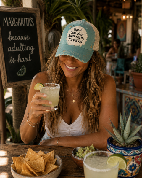 Woman wearing the BohoGroove Today’s Good Mood Sponsored By Margaritas hat with black lettering on a distressed off-white patch on an ocean blue vintage distressed snapback while enjoying a margarita on a colorful outdoor patio with chips and guacamole.