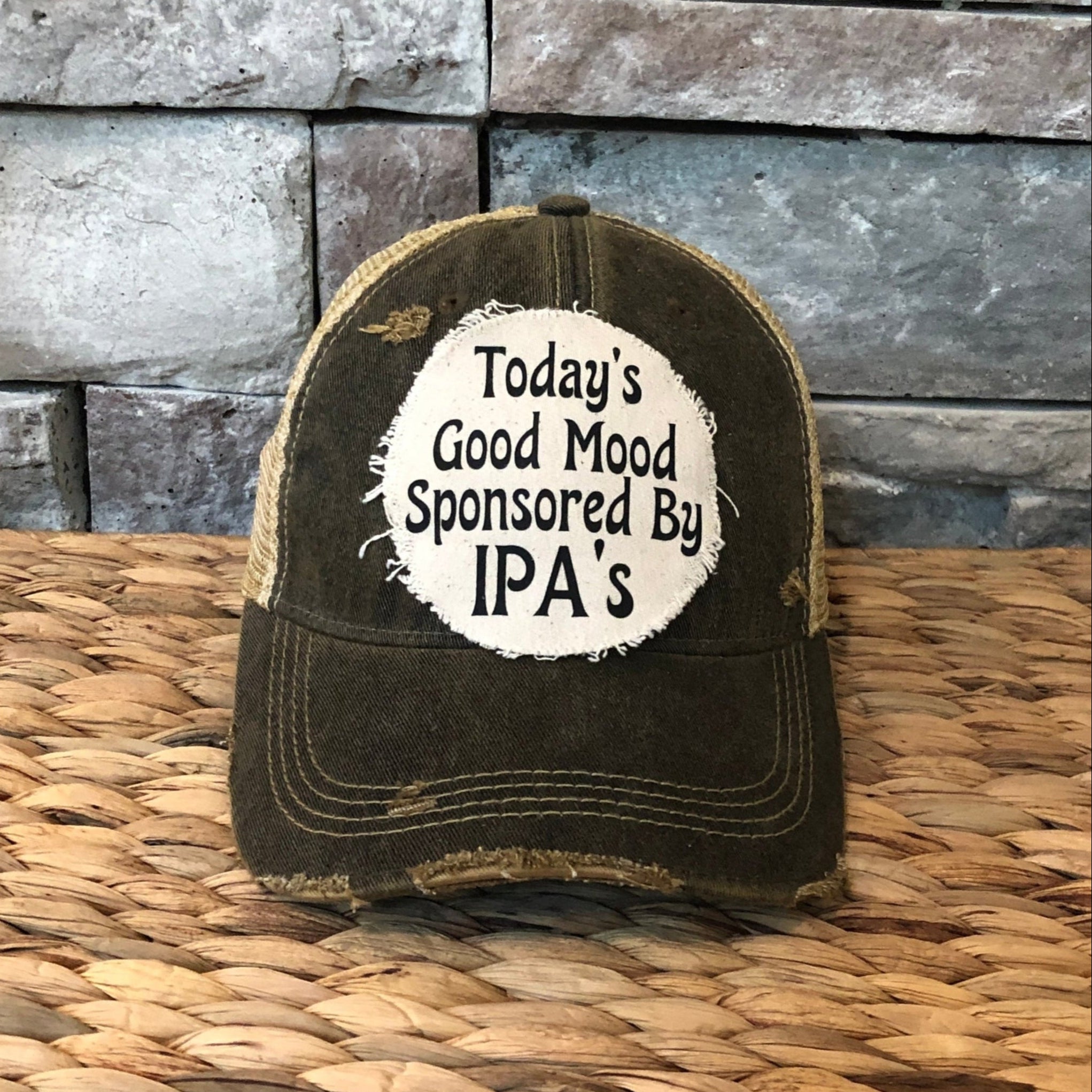 Distressed cap with 'Today's Good Mood Sponsored By IPA's' text on a woven surface and stone wall background