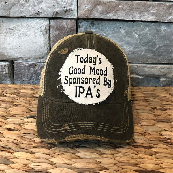 Distressed cap with 'Today's Good Mood Sponsored By IPA's' text on a woven surface and stone wall background