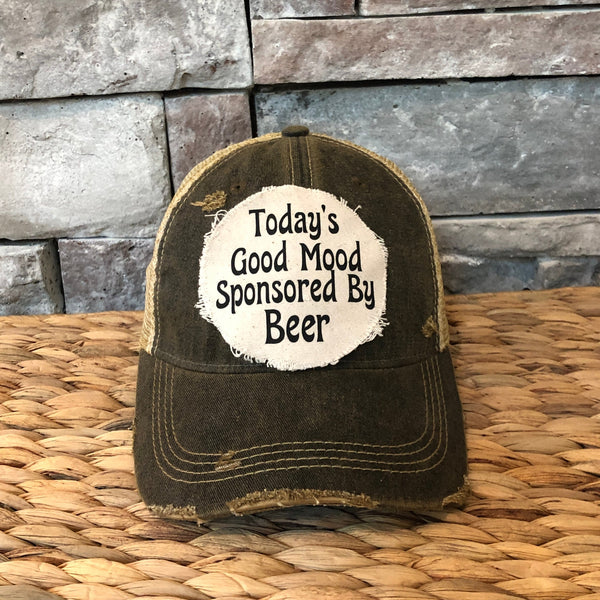 Distressed black cap with 'Today's Good Mood Sponsored By Beer' text on a woven surface and stone wall background.