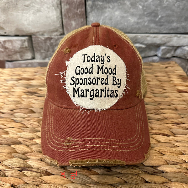 Brown cap with 'Today's Good Mood Sponsored By Margaritas' text on a woven surface and stone wall background