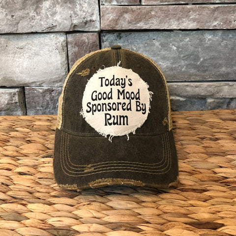 Brown distressed cap with 'Today's Good Mood Sponsored By Rum' text on a woven surface and stone wall background.