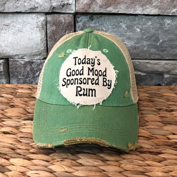 Green cap with text 'Today's Good Mood Sponsored By Rum' on a woven surface and stone wall background