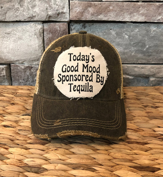 Distressed cap with text 'Today's Good Mood Sponsored By Tequila' on a woven surface and stone wall background