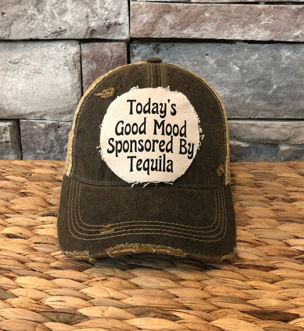 Distressed cap with text 'Today's Good Mood Sponsored By Tequila' on a woven surface and stone wall background