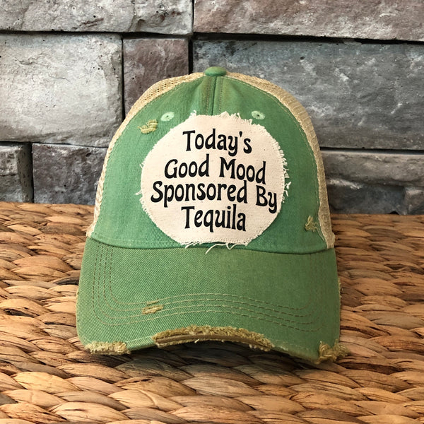 Green cap with text 'Today's Good Mood Sponsored By Tequila' on a woven surface and stone wall background