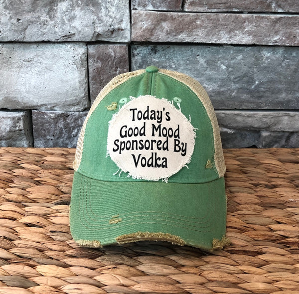 Green cap with text 'Today's Good Mood Sponsored By Vodka' on a woven surface and stone wall background