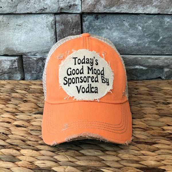 Orange cap with text 'Today's Good Mood Sponsored By Vodka' on a woven surface and stone wall background