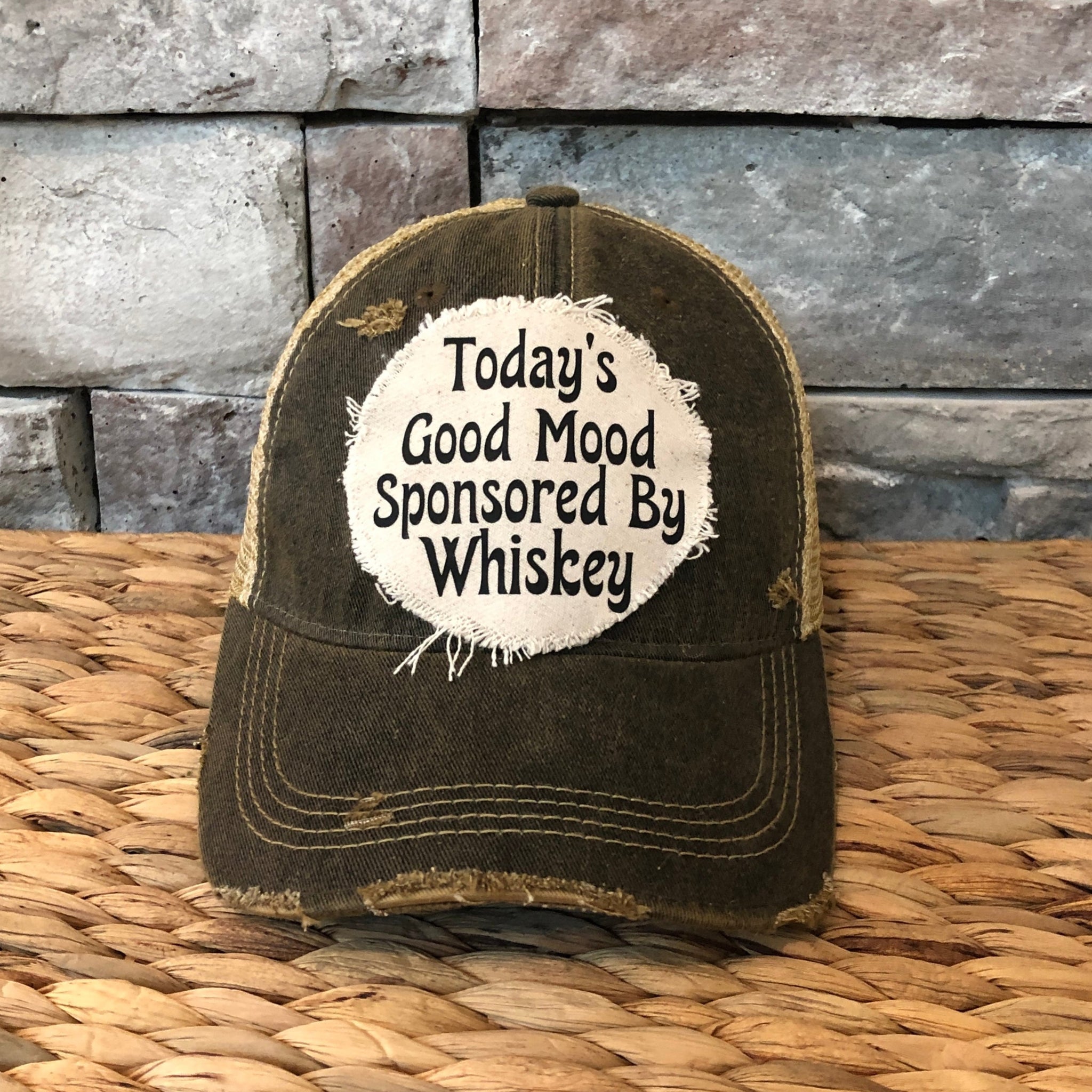Distressed cap with 'Today's Good Mood Sponsored By Whiskey' text on a woven surface with stone wall background