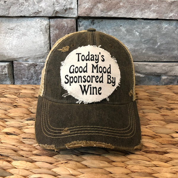 Black Distressed cap with 'Today's Good Mood Sponsored By Wine' text on a woven surface and stone wall background.