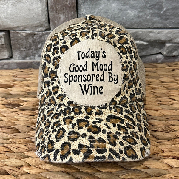 Leopard print cap with 'Today's Good Mood Sponsored By Wine' text on a woven surface and stone wall background
