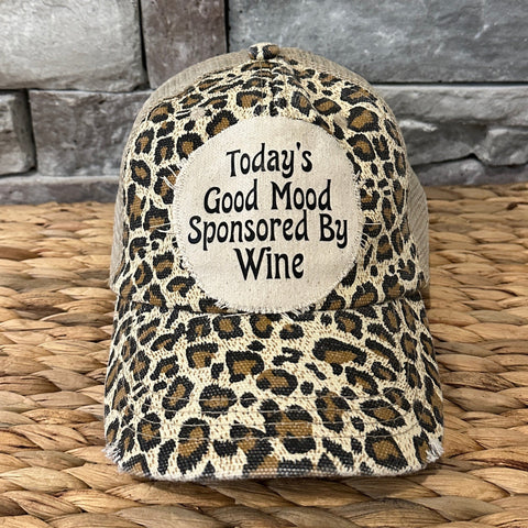 Leopard print cap with 'Today's Good Mood Sponsored By Wine' text on a woven surface and stone wall background