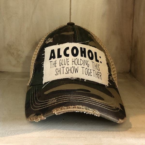 Alcohol: The Glue Holding This Shitshow Together hat by BohoGroove with black lettering on a distressed off-white patch on a camo vintage distressed snapback trucker hat.