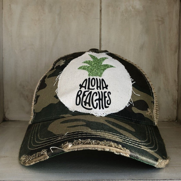 Aloha Beaches hat by BohoGroove with black Aloha Beaches lettering and green pineapple graphic on distressed off-white patch and camo vintage distressed snapback.