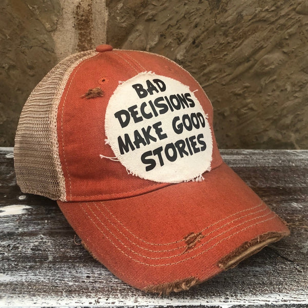 Left side view of Bad Decisions Make Good Stories hat by BohoGroove with black Bad Decisions Make Good Stories lettering on a distressed off-white patch on an orange vintage distressed snapback trucker hat.