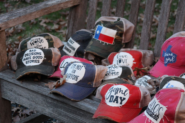 BohoGroove collection of vintage distressed snapback trucker hats with funny sayings, wine hats, game day hats, Texas hats, and custom patch designs displayed on a rustic wooden bench.