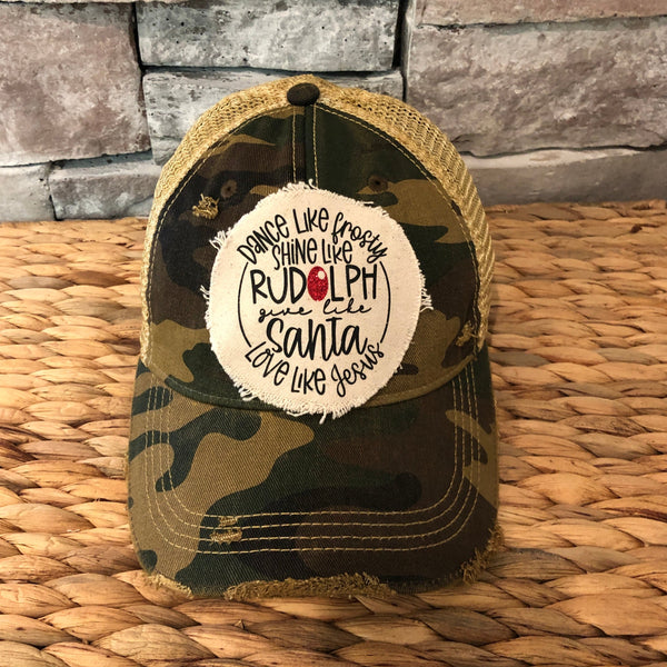Dance Like Frosty Shine Like Rudolph Give Like Santa Love Like Jesus hat by BohoGroove with red and green lettering and Christmas phrase on a distressed off-white patch on a camo vintage distressed snapback.