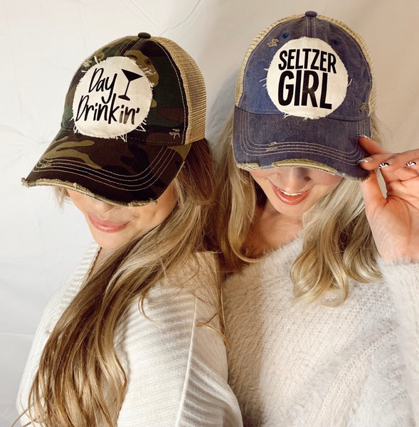 Two women wearing BohoGroove vintage distressed snapback trucker hats, featuring the Day Drinkin' hat on a camo hat and the Seltzer Girl hat on a navy blue hat with distressed off-white patches.