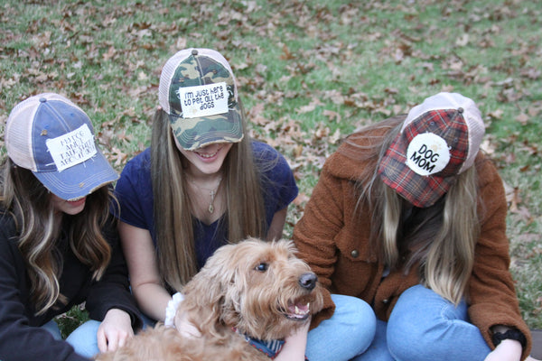 Women wearing BohoGroove dog-themed vintage distressed snapback hats including My Dog and I Talk Shit About You, I'm Just Here to Pet All the Dogs, and Dog Mom while sitting outdoors with a dog.