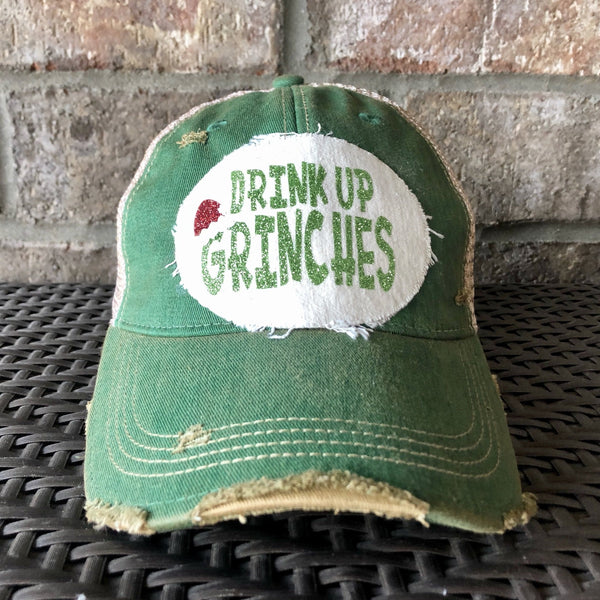 Drink Up Grinches hat by BohoGroove with glitter green Drink Up Grinches lettering and a red Santa hat graphic on a distressed off-white custom patch on a green vintage distressed snapback.