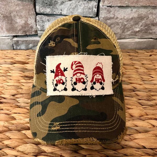 Gnome Ho Ho Ho hat by BohoGroove with three Christmas gnomes wearing red glitter hats and Ho Ho Ho lettering on a distressed off-white patch on a camo vintage distressed snapback.