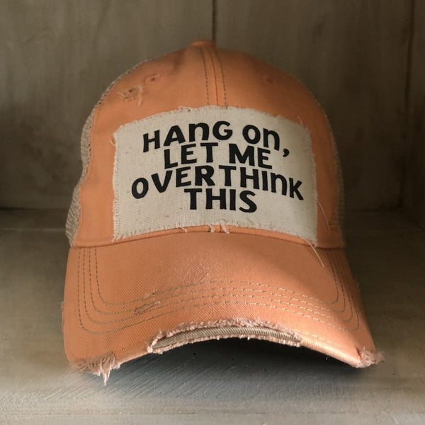 Hang On Let Me Overthink This hat by BohoGroove with black Hang On Let Me Overthink This lettering on a distressed off-white patch on a sherbet vintage distressed snapback trucker hat.