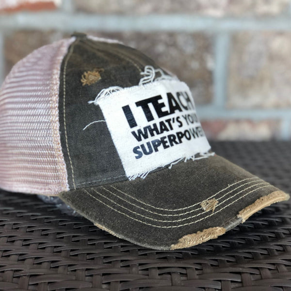 Left side view of I Teach What's Your Superpower hat by BohoGroove with black lettering on a distressed off-white patch on a black vintage distressed snapback trucker hat.