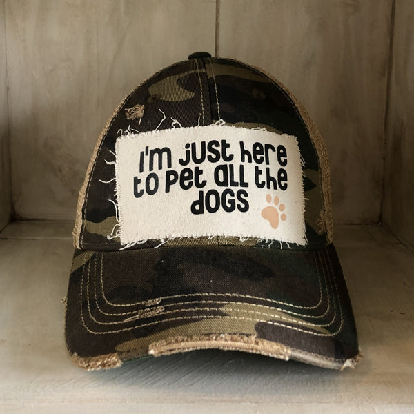 I'm Just Here to Pet All the Dogs hat by BohoGroove with black I'm Just Here to Pet All the Dogs lettering and paw print graphic on a distressed off-white patch on a camo vintage distressed snapback.